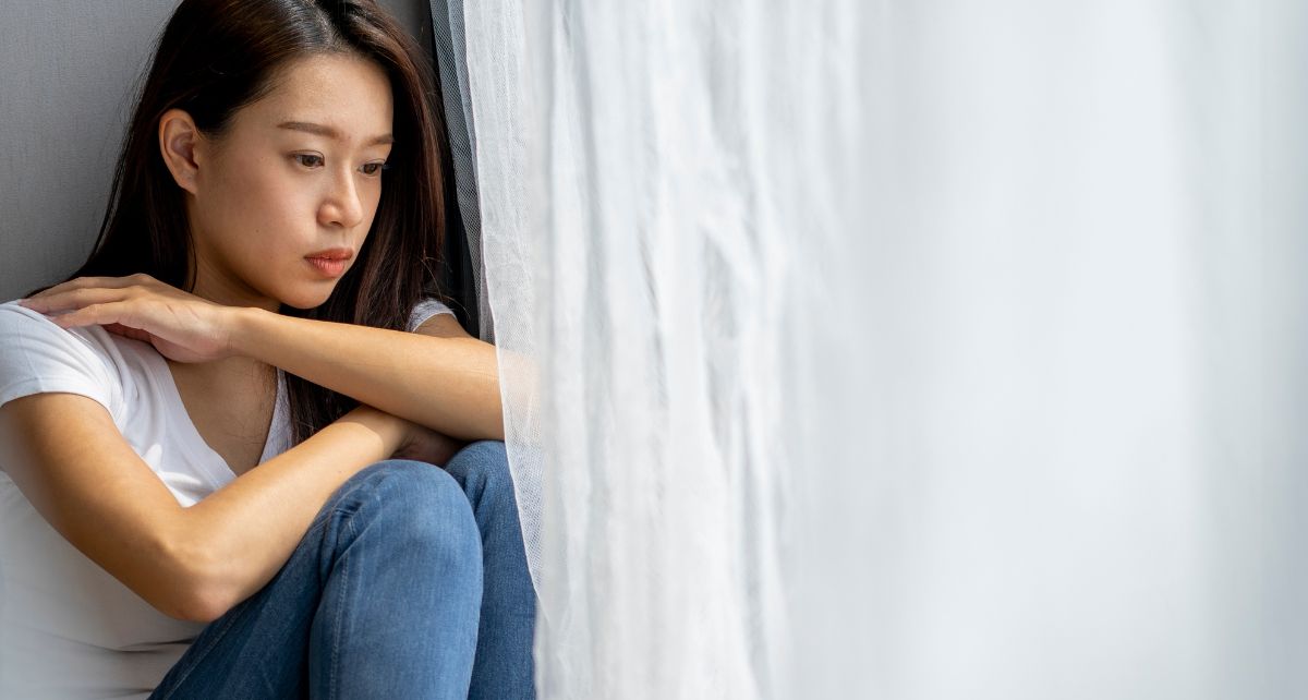 Woman looks out a window with white curtains struggling with PMDD