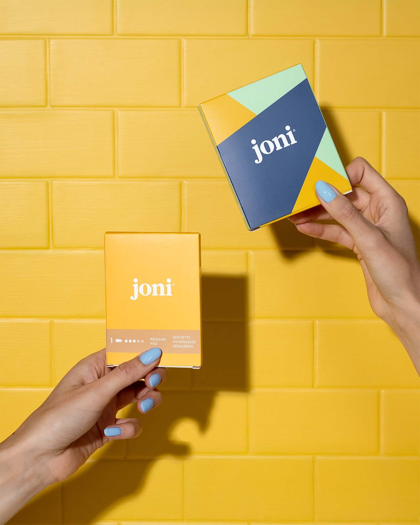Two 'joni' product boxes held against a yellow tiled wall.