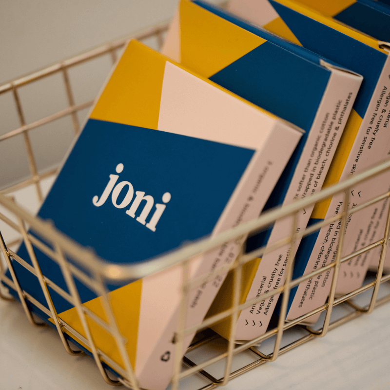 Colorful boxes of joni period care products arranged in a wire basket, promoting period equity.