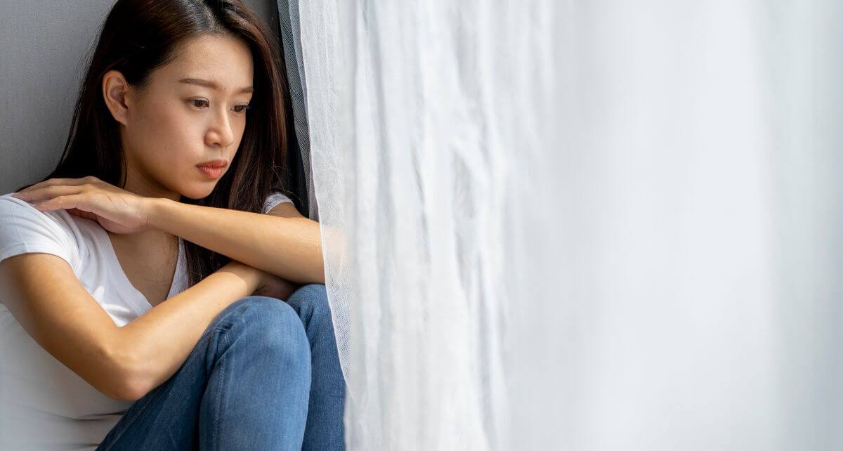 Woman looks out a window with white curtains struggling with PMDD
