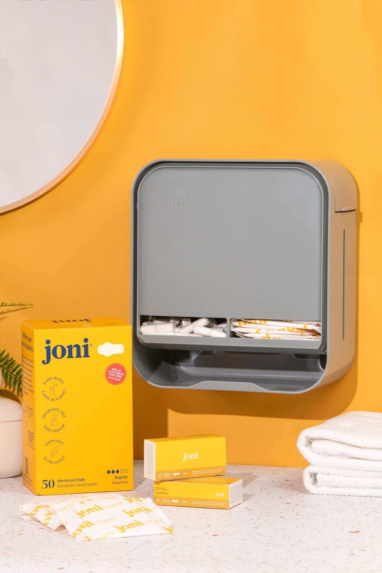 Modern joni open period care dispenser on a countertop with pads and tampons, set against a vibrant yellow wall.