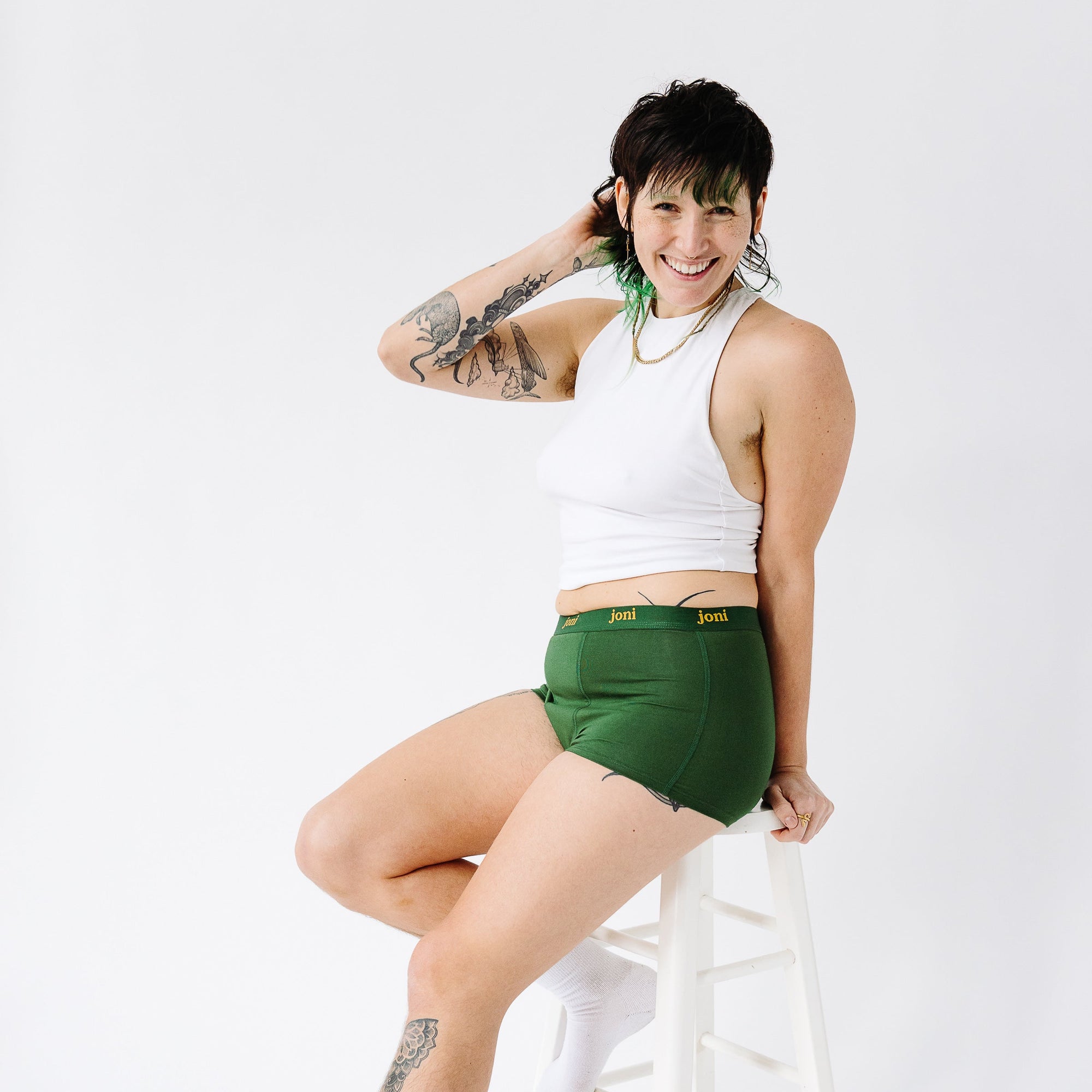 Person in joni green period underwear sits on a stool wearing a white tank top