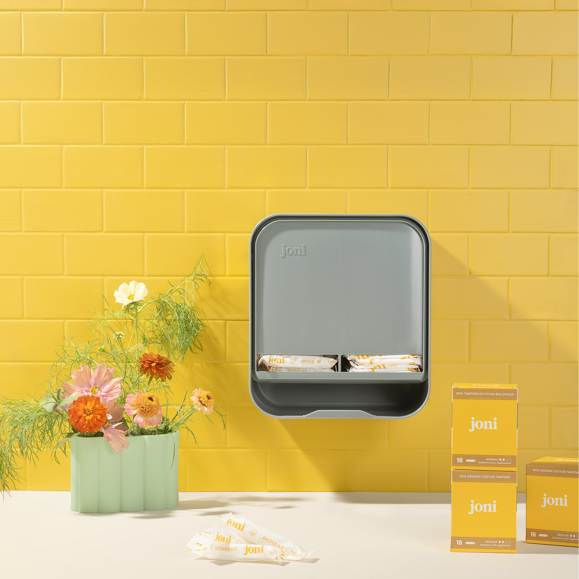 joni Model A open period care holder hanging on a yellow brick wall with tampons and flowers on the bathroom counterop