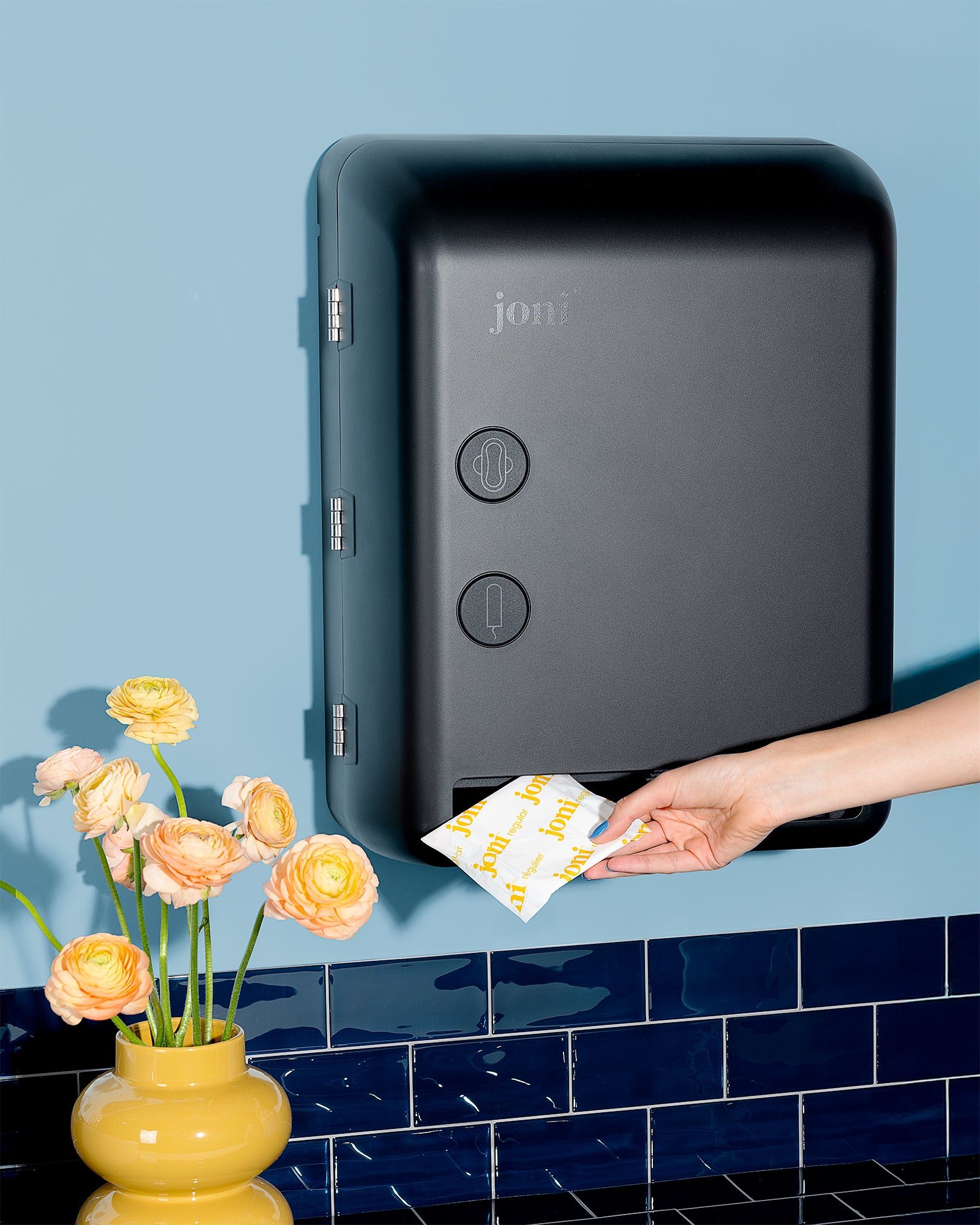 Hand placing a packet into a joni period care dispenser against a blue tiled wall with flowers in a workplace washroom.