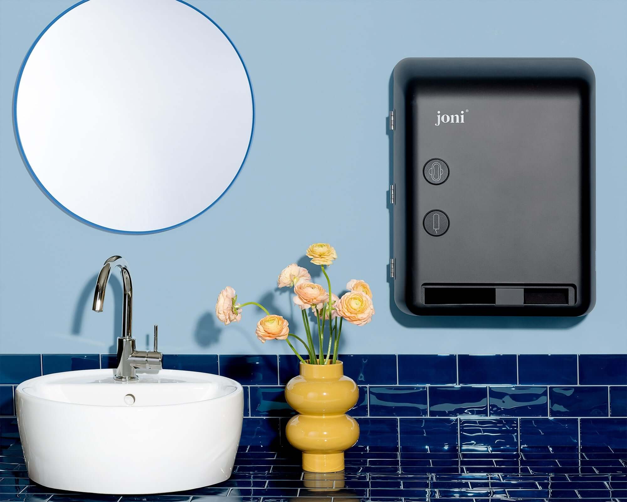 Bathroom setting with a sink, mirror, and decorative vase with the joni model v dispenser hanging on a blue tiled wall