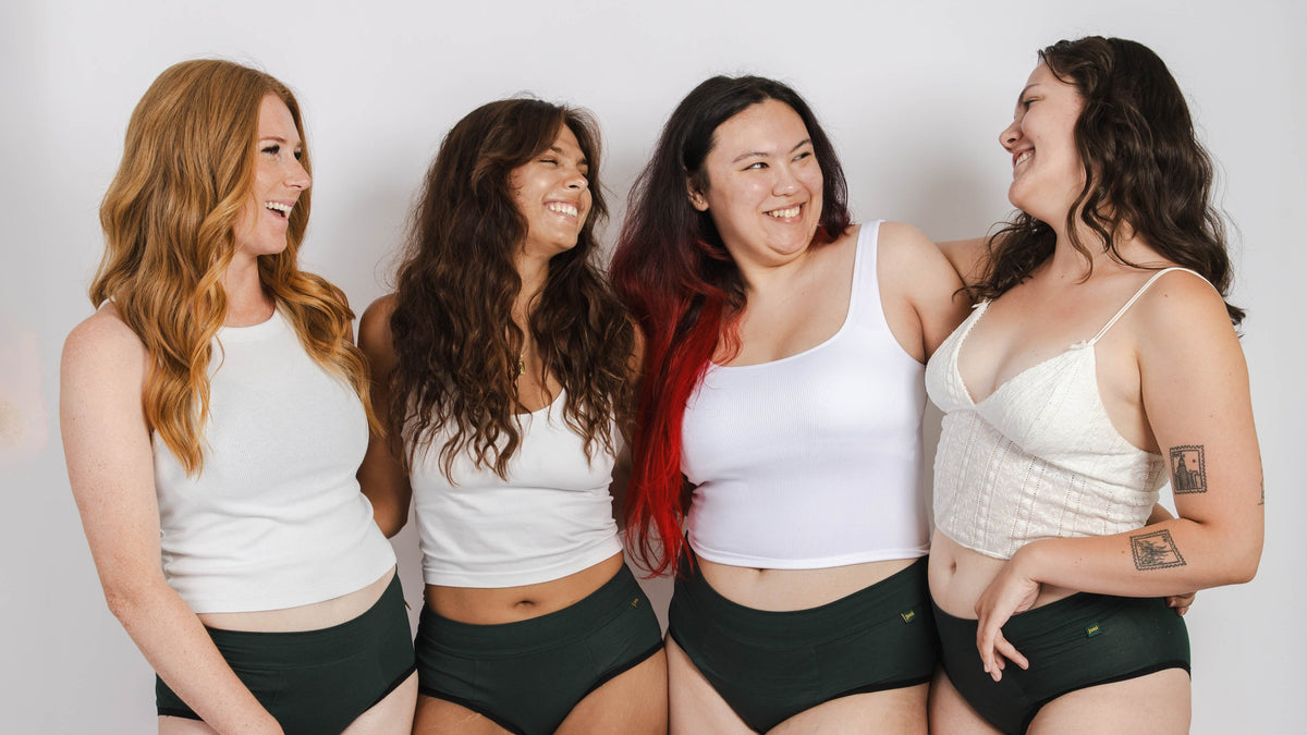 Four women standing together in a studio wearing matching white tank tops, joni period underwear, white socks, and white underwear.